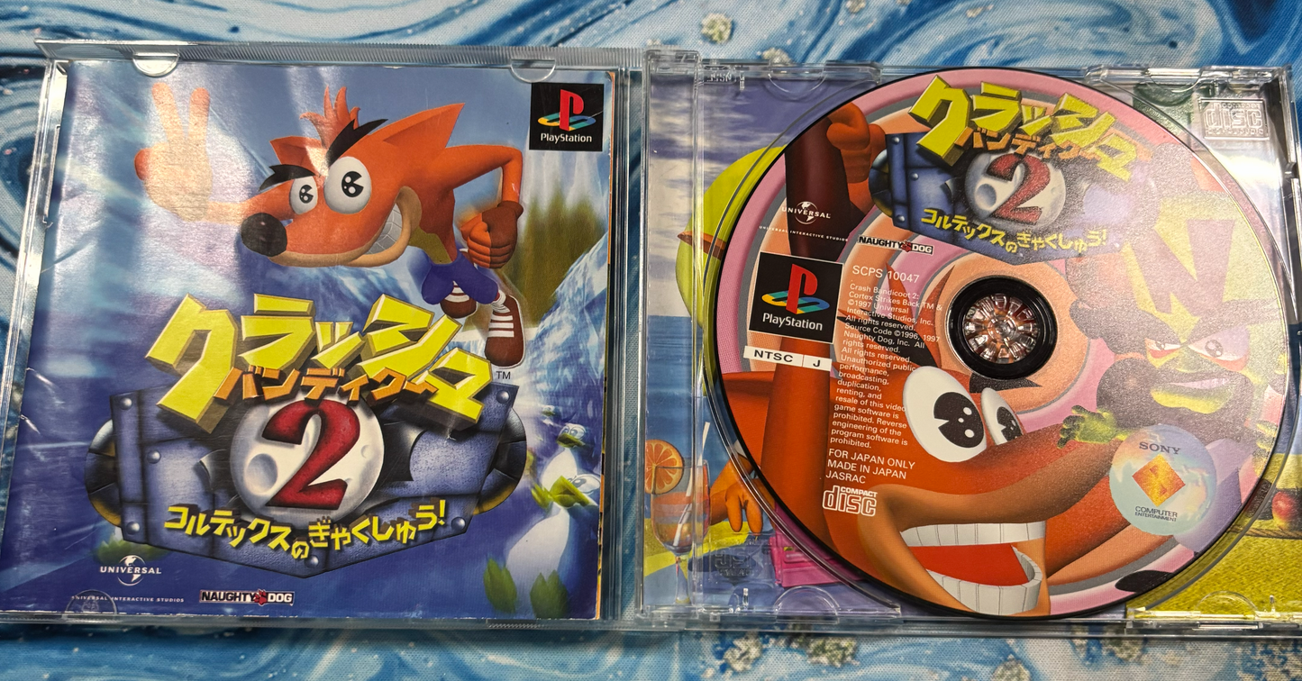 A PlayStation 1 game case for Crash Bandicoot 2:howing both the front cover art and the printed disc on a blue patterned surface.