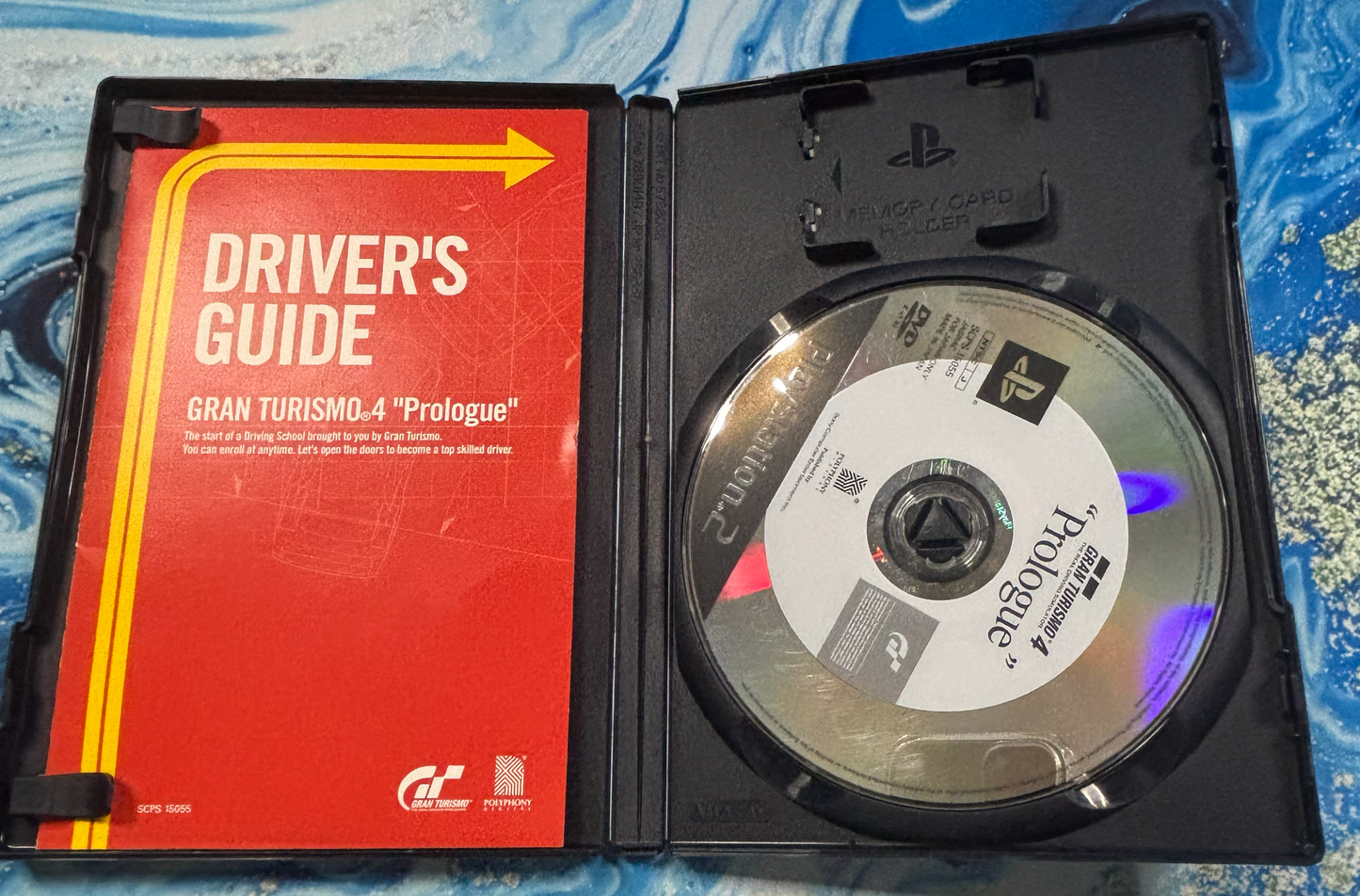 Open PlayStation 2 game case for Gran Turismo 4 'Prologue' showing the red and yellow driver's guide insert and the game disc on a blue patterned surface.