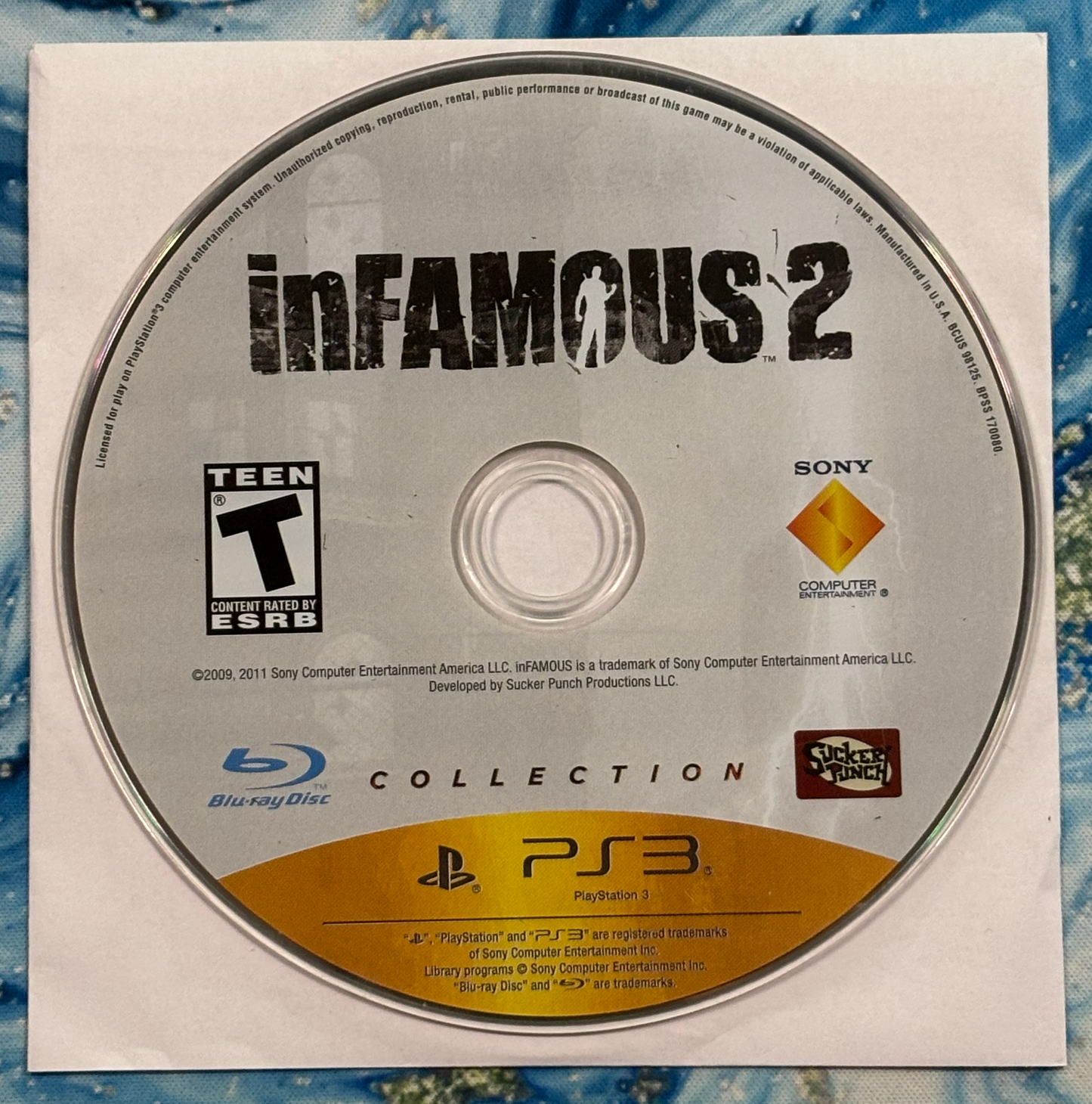 Game disc for 'inFAMOUS 2' on a blue textured background