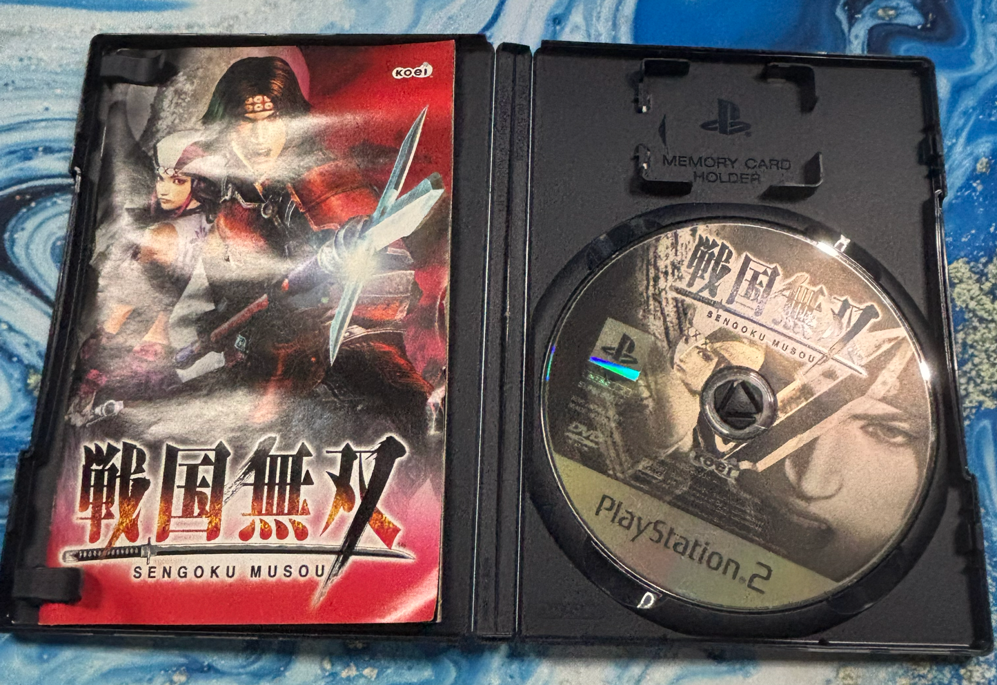 Open PlayStation 2 game case for Sengoku Musou, showing the printed game cover on the left and the game disc on the right, placed on a blue patterned fabric background.