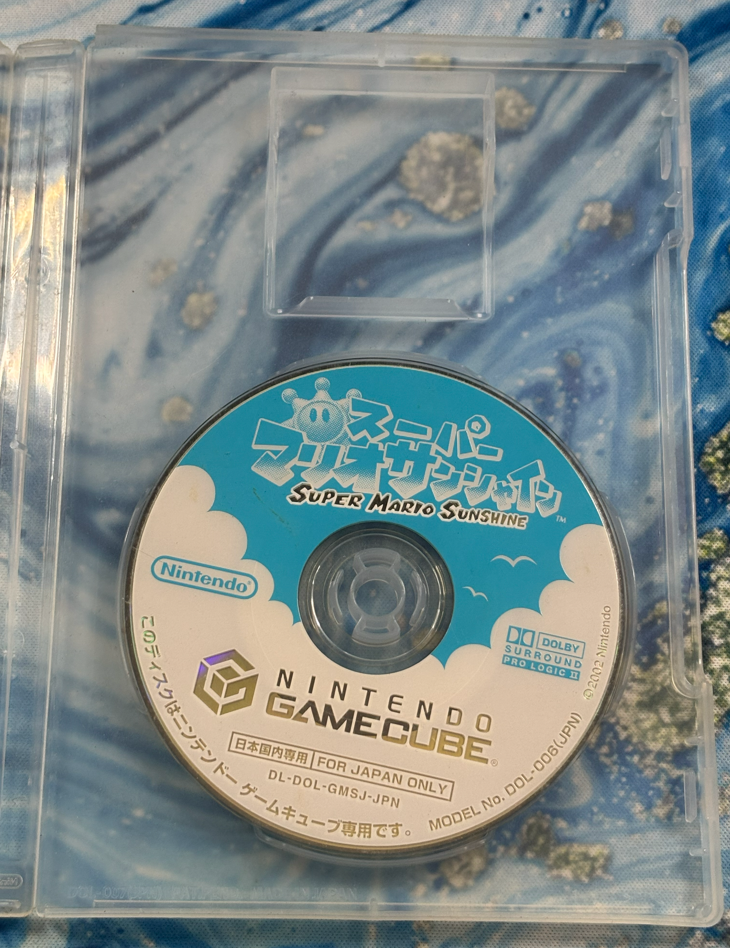 Super Mario Sunshine game disc for Nintendo GameCube, displayed inside a clear plastic jewel case against a blue and white marble-patterned background.