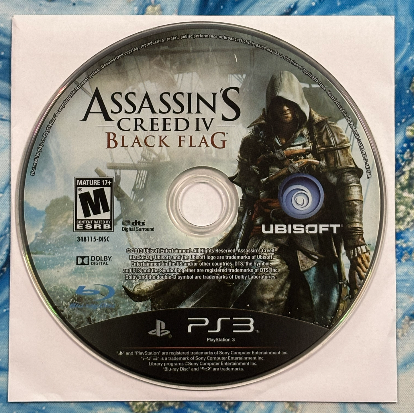 Assassin's Creed IV Black Flag game disc for PlayStation 3 on a blue and white patterned background