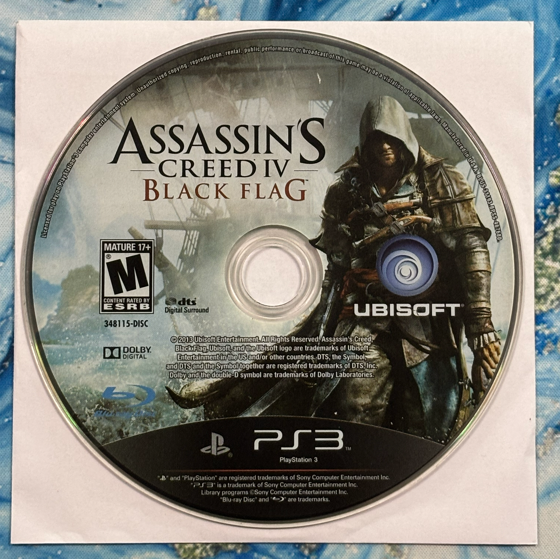 Assassin's Creed IV Black Flag game disc for PlayStation 3 on a blue and white patterned background