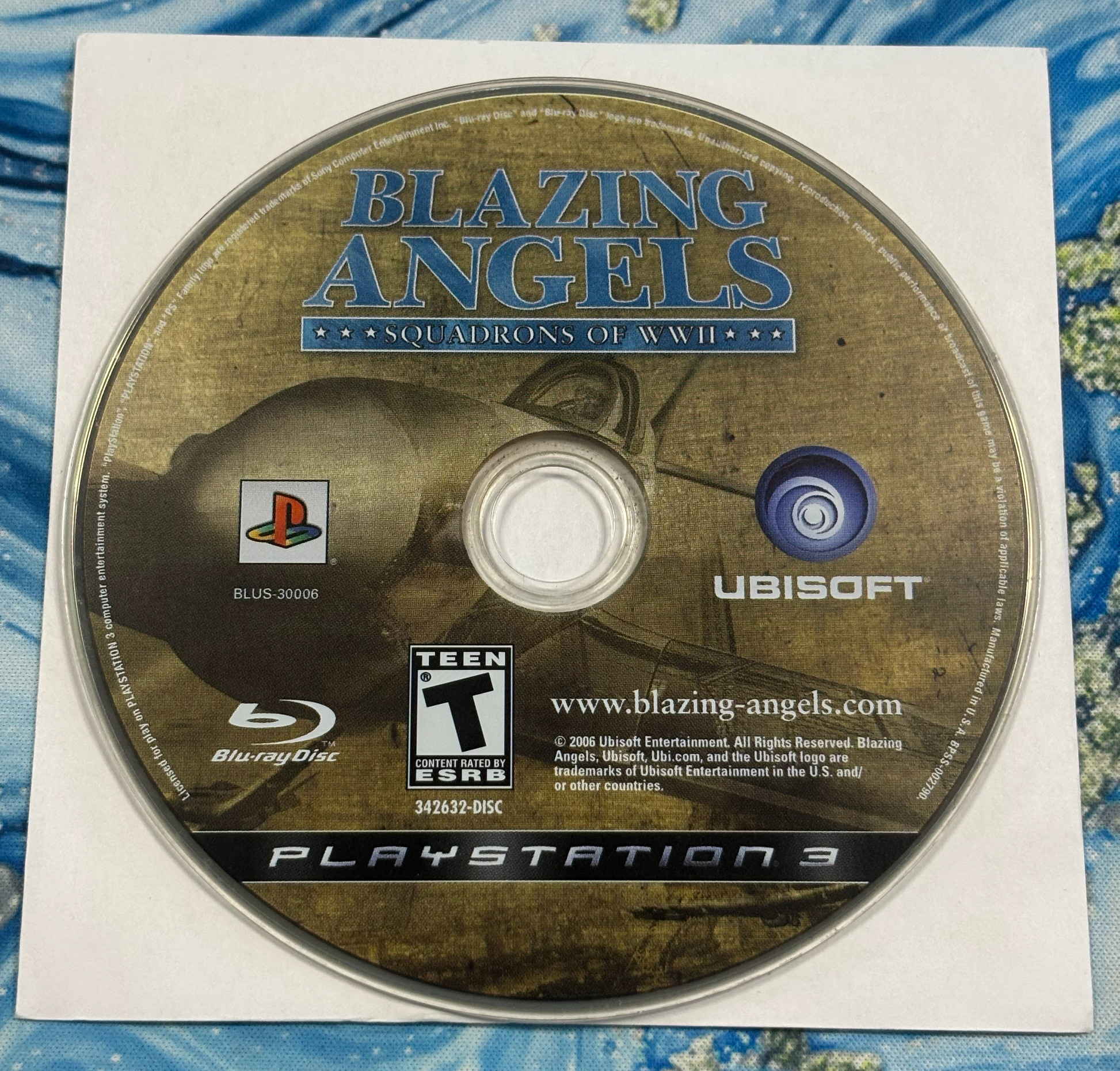A PlayStation 3 Blu-ray game disc for the video game Blazing Angels: Squadron of War II, shown on its white paper sleeve against a blue patterned fabric background.
