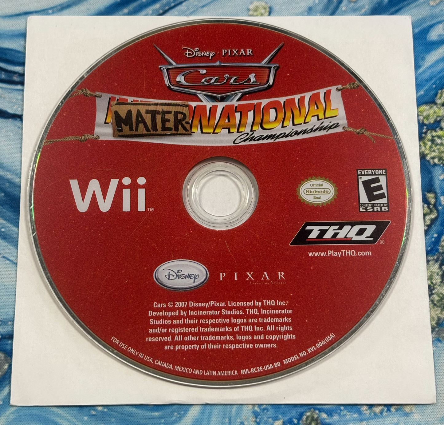 A used Disney Pixar Cars: Mater National Championship video game disc for the Nintendo Wii, still in its original white paper sleeve, placed on a blue patterned surface.