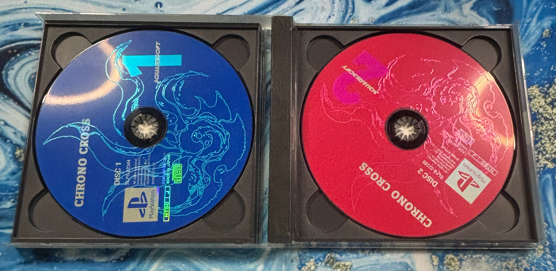 Two PlayStation 1 game discs in black jewel cases, one blue with Chroma Cross artwork and one red with a different cover design, placed on a blue patterned surface.