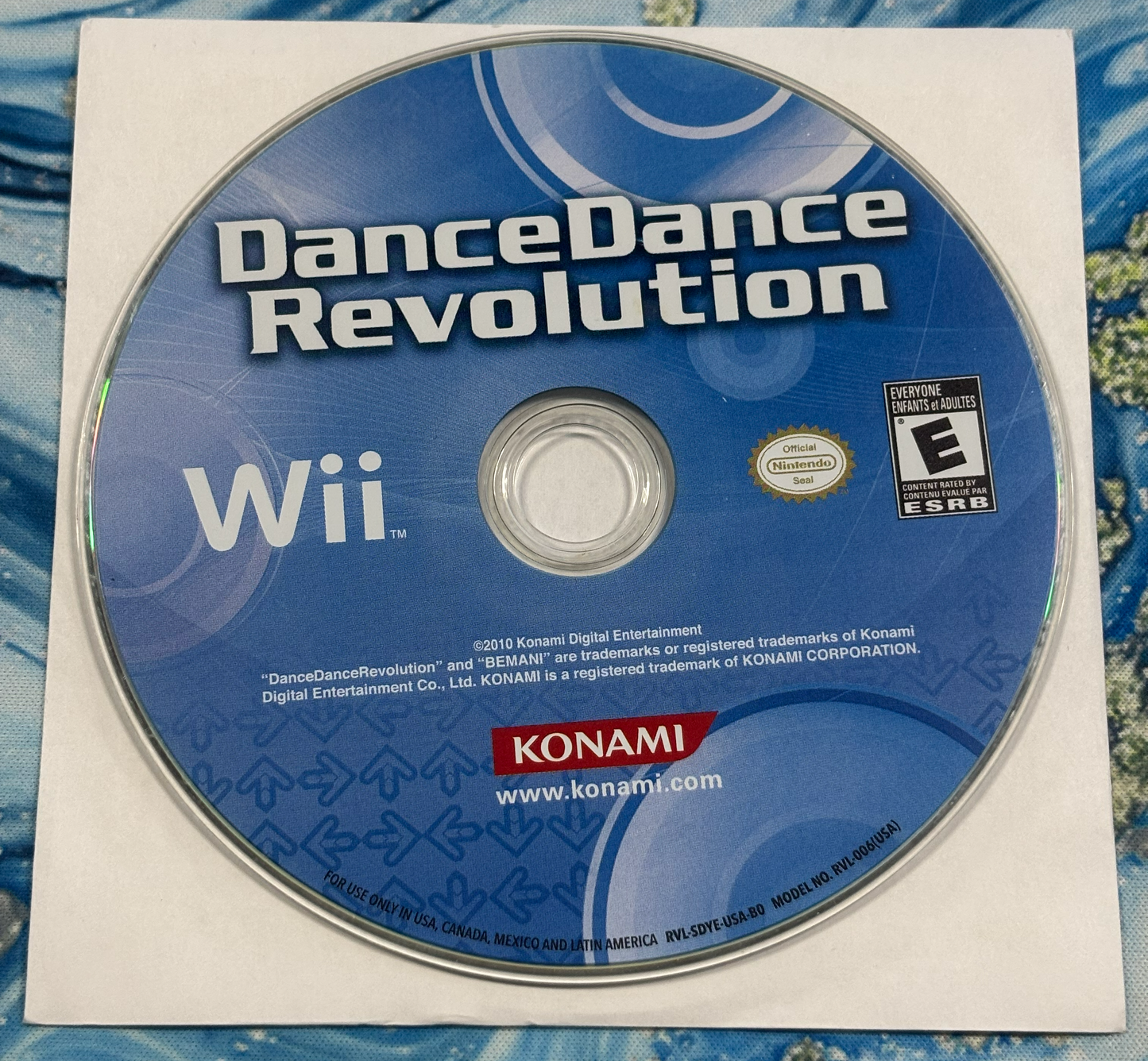 A used Nintendo Wii game disc for DanceDance Revolution, still in its original white paper sleeve, photographed flat on a blue patterned fabric background.