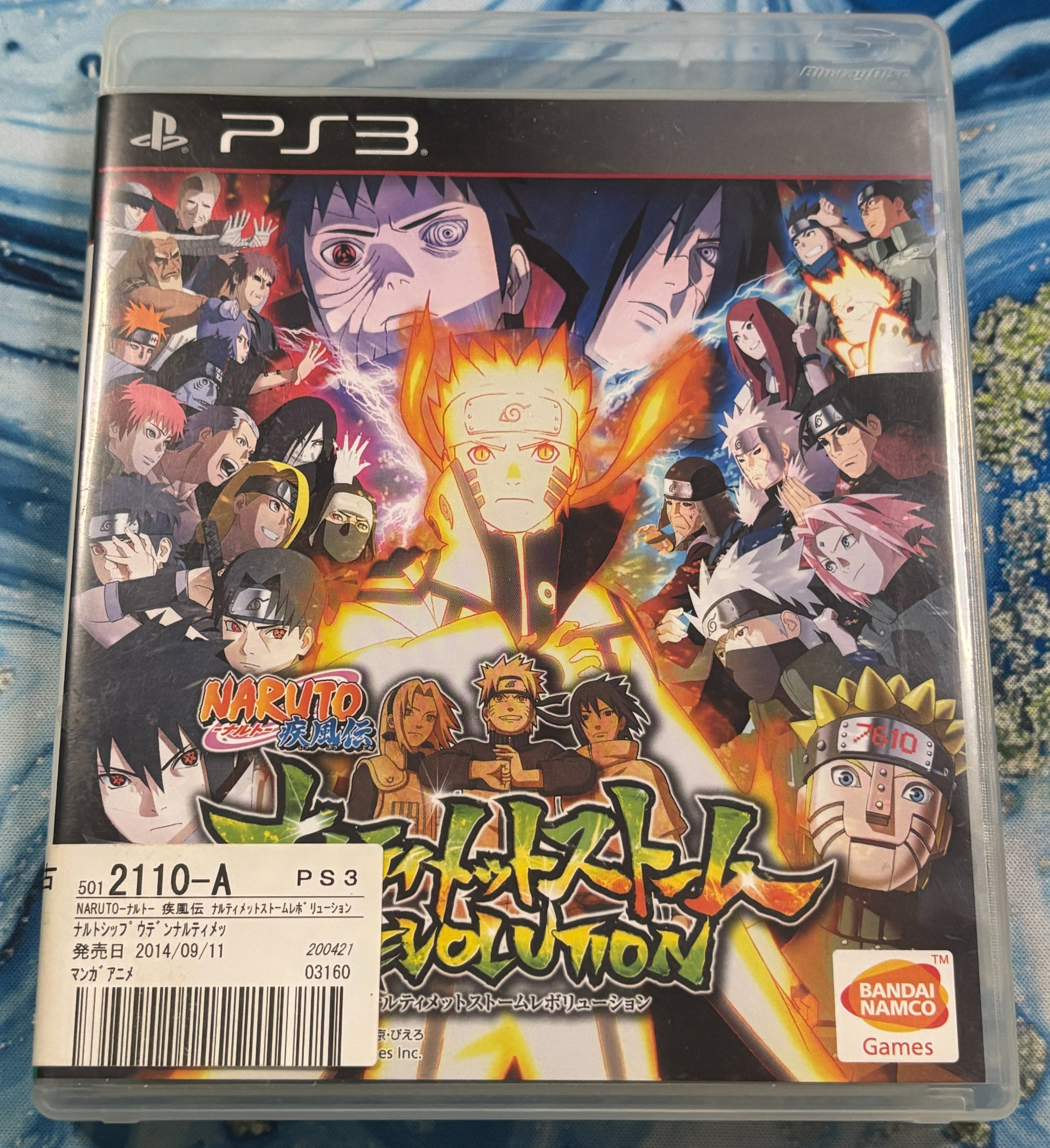 PS3 game case for Naruto Shippuden: Ultimate Volume 10, featuring a colorful anime character illustration on the cover, photographed in a clear plastic case on a blue patterned fabric background.