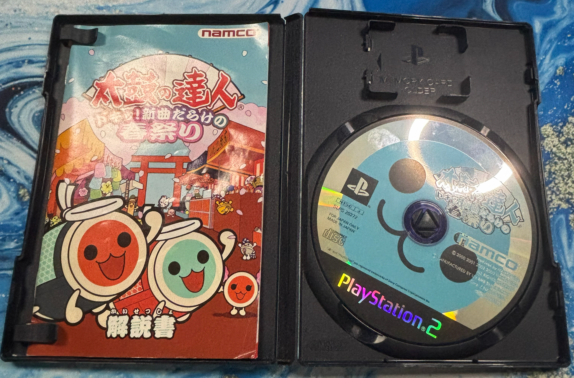 Open PlayStation 2 game case showing the Japanese cover art and disc for Namco's Toad Meme Toad Taisen 2 game, placed on a blue patterned fabric background.